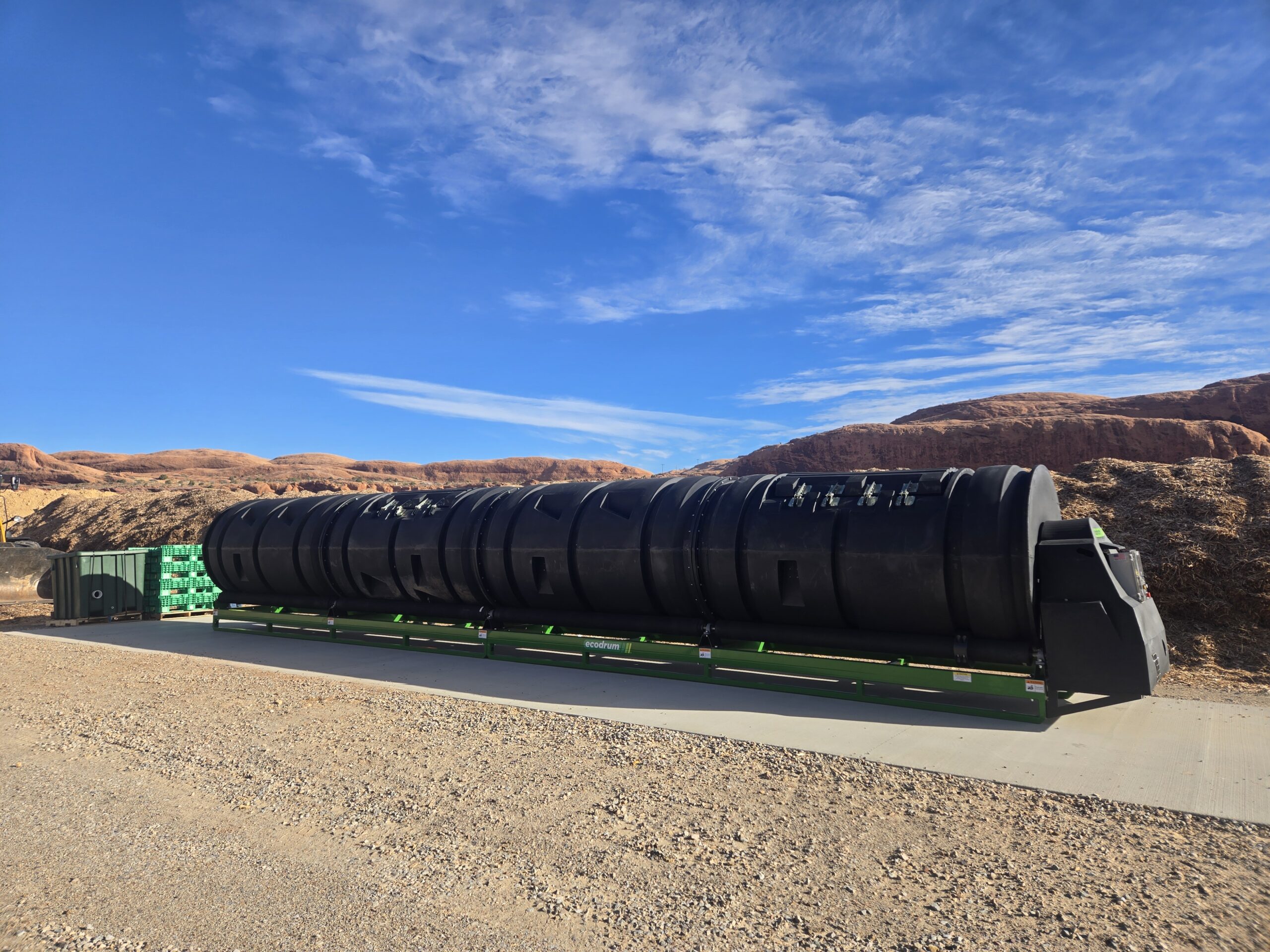 A large black cylindrical structure on a concrete pad in a desert landscape. Blue sky with clouds and reddish-brown hills in the background.